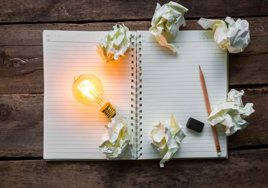 A lit light bulb on an open notebook surrounded by paper ideas